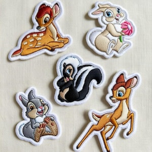 Può includere: Set di cinque toppe ricamate con personaggi di un classico film d'animazione. Include un cerbiatto, un coniglio con un fiore, una puzzola e altri due cervi. Ogni toppa ha un bordo bianco ed è multicolore.