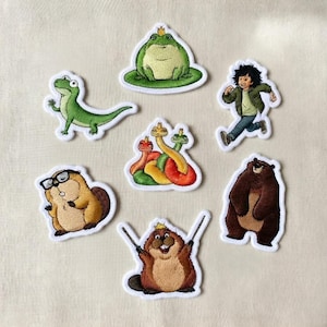 May include: A collection of embroidered patches featuring various cartoon characters. The patches include a green frog with a crown, a running boy, a brown bear, a lizard, a beaver wearing glasses, a beaver with a crown, and colorful snakes.