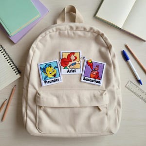 May include: Beige backpack with three embroidered patches featuring characters from The Little Mermaid: Flounder, Ariel, and Sebastian. The backpack has a front pocket and a top handle. The patches are rectangular with white borders.