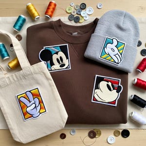 May include: A brown sweatshirt, gray beanie, and canvas tote bag with Mickey Mouse and hand gesture patches. The items are surrounded by spools of thread and buttons, suggesting a craft or DIY theme.