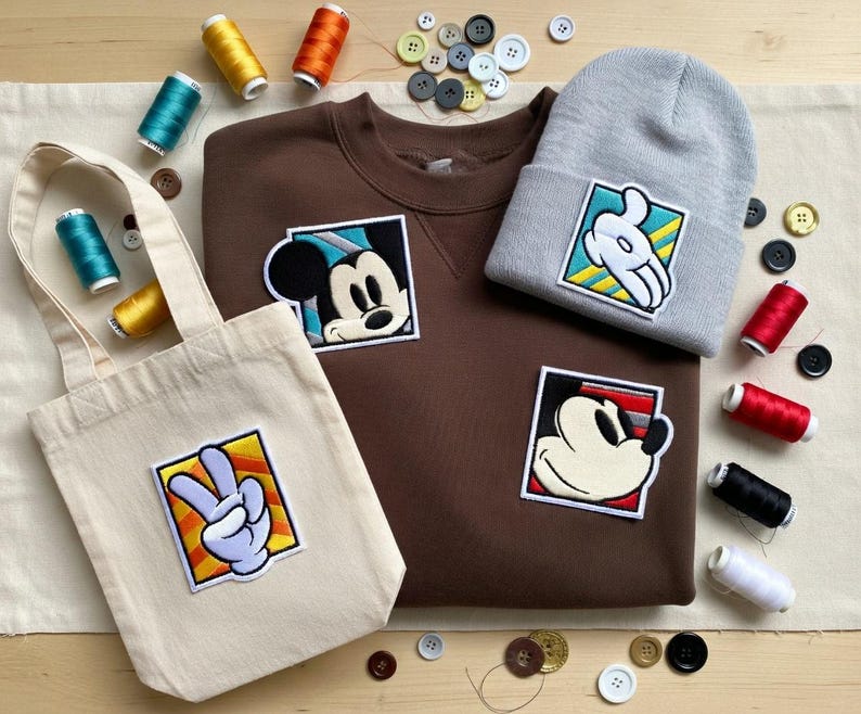 May include: A brown sweatshirt, gray beanie, and canvas tote bag with Mickey Mouse and hand gesture patches. The items are surrounded by spools of thread and buttons, suggesting a craft or DIY theme.