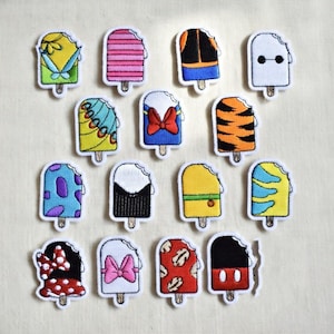 Puede incluir: Una colección de parches bordados con forma de helado, coloridos y con diversos diseños, incluyendo rayas y personajes. Cada parche tiene un borde blanco y un palito de madera, ideal para decorar ropa o accesorios.