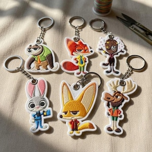 May include: A collection of six embroidered character keychains featuring various anthropomorphic animals, including a sloth, a fox, a yak, a rabbit, a fennec fox, and a moose, each attached to a silver-toned metal split ring for keys.