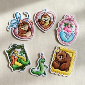 May include: Embroidered patches featuring various animals, including a bear, beaver, and lizard, each wearing a party hat. The patches are in different shapes, such as hearts and frames, and are outlined in white.