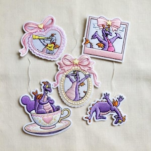 May include: Set of five embroidered patches featuring a purple dragon character in various poses. Each patch has a different shape, including a heart, a square, and an oval, and is decorated with pink bows and accents. The patches are on a cream-colored background.