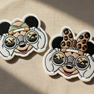 May include: Two embroidered patches featuring Mickey and Minnie Mouse with binoculars. Mickey wears a safari hat, while Minnie has a bow with leopard spots. Both are looking through binoculars at a savanna scene.