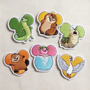 May include: Set of six embroidered patches featuring whimsical cartoon animals. Designs include a green lizard, a brown beaver with a crown, a caterpillar, a pink creature in a bowl, a brown beaver, and a white goose with a yellow heart.