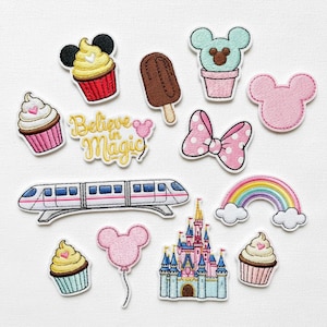 Può includere: Una collezione di toppe ricamate con vari disegni. Include cupcakes, un gelato, un fiocco, un arcobaleno, un treno, un castello e un palloncino. È presente anche il testo "Believe in Magic".