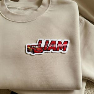 Puede incluir: Sudadera beige con un parche rojo y blanco con el nombre "LIAM" y un coche de carreras de dibujos animados. El parche est&aacute; cosido en la parte delantera de la sudadera.