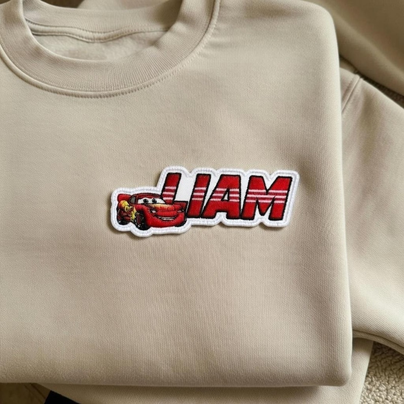 Puede incluir: Sudadera beige con un parche rojo y blanco con el nombre "LIAM" y un coche de carreras de dibujos animados. El parche est&aacute; cosido en la parte delantera de la sudadera.