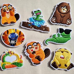 May include: A collection of embroidered patches featuring various cartoon animals. The patches include a brown bear, a green lizard wearing sunglasses, and several beaver-like creatures in shades of orange and yellow. Other patches include a frog and a creature with a blue face.