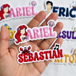 May include: Embroidered patches featuring characters and names from a popular animated film. The patches include the names "Ariel" in pink and "Sebastian" in red, along with character illustrations and related icons. The patches are on a white background.