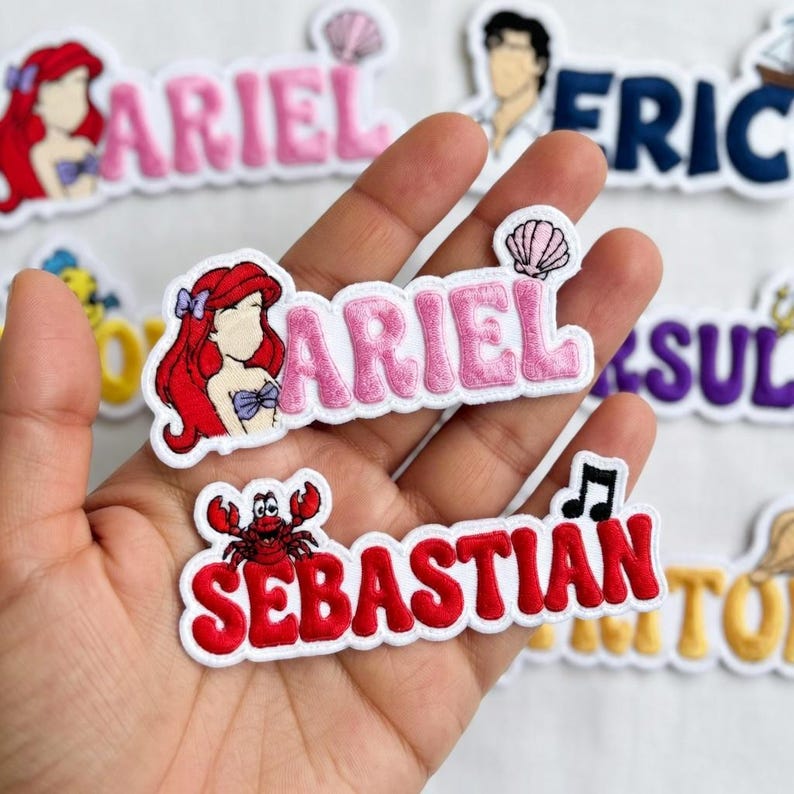 May include: Embroidered patches featuring characters and names from a popular animated film. The patches include the names "Ariel" in pink and "Sebastian" in red, along with character illustrations and related icons. The patches are on a white background.