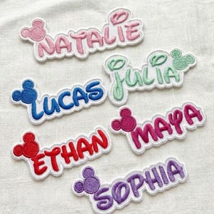 May include: Embroidered name patches in various colors, including pink, blue, red, green, purple, and magenta. Each patch features a name with a Mickey Mouse head design. Names include Natalie, Lucas, Ethan, Julia, Maya, and Sophia.