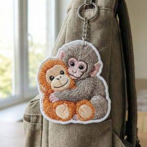 May include: Embroidered keychain featuring two hugging monkeys. One monkey is brown, the other is gray, both with smiling faces. The keychain is attached to a silver chain and a metal ring, hanging from a tan backpack.