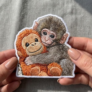 Op de afbeelding: Geborduurde patch met twee knuffelende apen. Eén aap is bruin, de andere oranje, beide met gedetailleerde steken. De patch heeft een witte rand. De patch wordt door een persoon vastgehouden.
