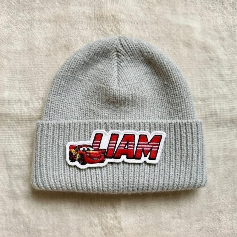 Puede incluir: Un gorro de punto gris con un borde doblado. El gorro presenta un parche con el nombre "LIAM" en letras rojas y un gr&aacute;fico de un coche de dibujos animados.