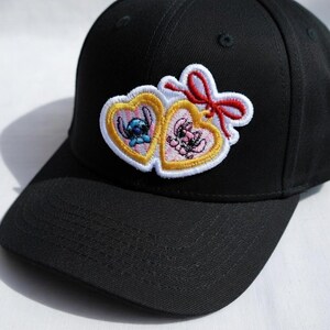 Op de afbeelding: Zwarte baseballcap met een geborduurde patch. De patch heeft twee hartvormige frames, elk met een cartoonfiguur, &eacute;&eacute;n blauw en &eacute;&eacute;n roze, en een rode strik. De cap is gemaakt van een stevige stof.