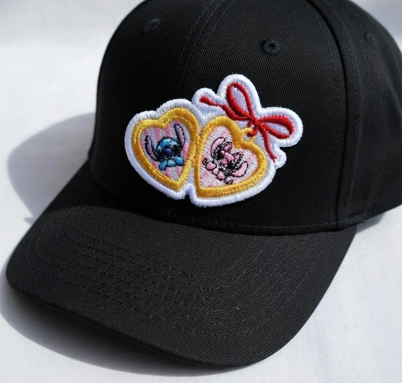 Op de afbeelding: Zwarte baseballcap met een geborduurde patch. De patch heeft twee hartvormige frames, elk met een cartoonfiguur, &eacute;&eacute;n blauw en &eacute;&eacute;n roze, en een rode strik. De cap is gemaakt van een stevige stof.