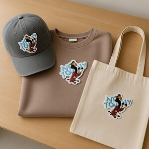 May include: A gray baseball cap, a brown sweatshirt, and a beige tote bag are displayed on a wooden surface. Each item features a colorful graphic of a cartoon character with the text "Not Today".