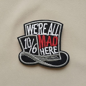 May include: Embroidered patch featuring a black top hat design with the text "WE'RE ALL MAD HERE." The word "MAD" is in red, and "10%" is on the left side. The patch has a white border and is on a beige background.