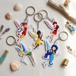 May include: Five embroidered fairy keychains featuring diverse figures with colorful wings and outfits. Each keychain includes a metal ring and chain attached to a fabric fairy shape. The designs include various hair colors, wing styles, and clothing.