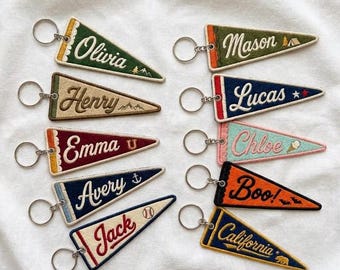 May include: Ten triangular felt pennant keychains with metal rings, each featuring embroidered names and small icons including mountains, an anchor, a baseball, a star, an ice cream cone, bats, and a bear in various colors like green, blue, red, and orange.