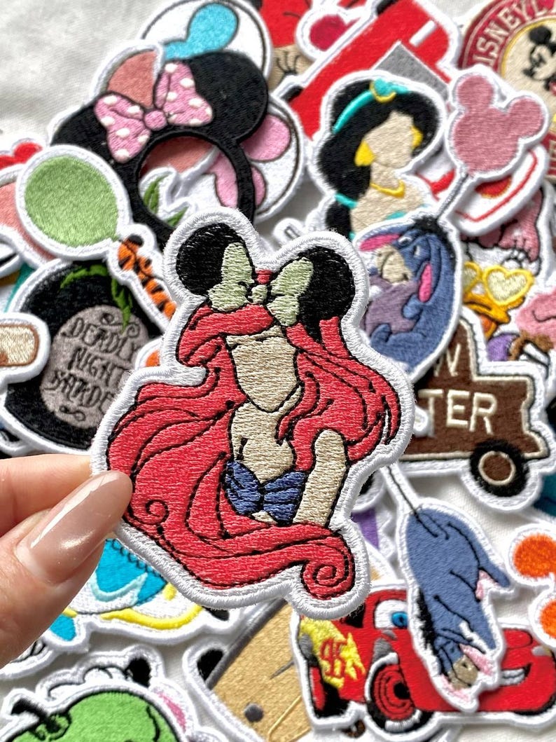 May include: A collection of colorful embroidered patches featuring various Disney characters. The patches include Ariel with red hair, Minnie Mouse with a pink bow, and Jasmine. The patches have white borders and are likely for clothing or accessories.