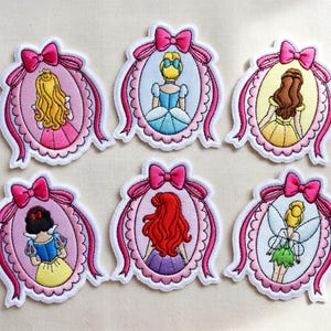 Puede incluir: Parches bordados con diseños de princesas. Cada parche presenta una princesa en un marco ovalado, adornado con un lazo y una cinta rosas. Los parches tienen un borde blanco y están sobre un fondo neutro.