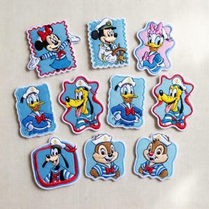 May include: Embroidered patches featuring Disney characters in sailor outfits. The collection includes Mickey Mouse, Minnie Mouse, Donald Duck, Daisy Duck, Goofy, Pluto, Chip, and Dale. Each patch has a blue background and a red border.