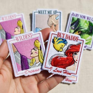 May include: Embroidered patches featuring cartoon characters and phrases. One patch shows a blonde character with the text "Wildest Dreams". Another patch shows a red-haired character with the text "But Daddy I Love Him". Other patches include the phrases "Meet Me At" and "Baby Now We Got".