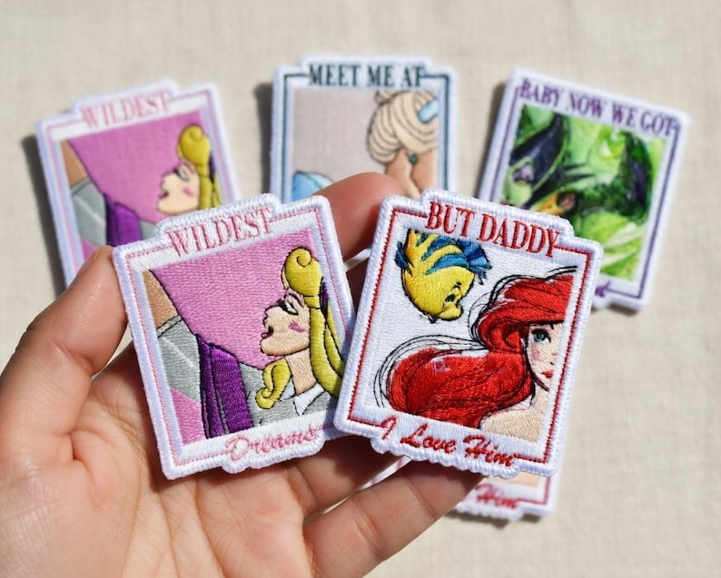 May include: Embroidered patches featuring cartoon characters and phrases. One patch shows a blonde character with the text "Wildest Dreams". Another patch shows a red-haired character with the text "But Daddy I Love Him". Other patches include the phrases "Meet Me At" and "Baby Now We Got".