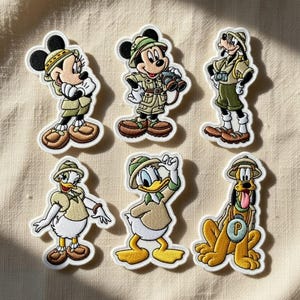 May include: A set of six embroidered patches featuring Disney characters dressed in safari gear. The collection includes Minnie Mouse, Mickey Mouse, Goofy, Daisy Duck, Donald Duck, and Pluto, all wearing khaki hats and outfits with brown accents.