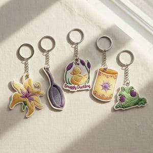 May include: Five embroidered keychains featuring a yellow flower, a purple spoon, a yellow duck with the text The Snuggly Duckling, a yellow lantern with a purple sun design, and a green frog with large purple eyes, all attached to silver rings.