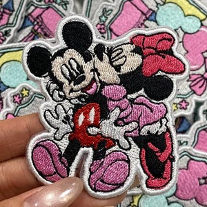 Puede incluir: Parche bordado con Mickey y Minnie Mouse en un abrazo amoroso. El parche tiene un borde blanco y muestra a los personajes icónicos en negro, rojo, rosa y blanco. El fondo incluye un castillo y elementos florales.