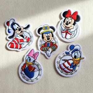 Puede incluir: Parches bordados con los personajes de Disney Mickey Mouse, Minnie Mouse, Goofy, Donald Duck y Daisy Duck. Cada parche está en un marco circular con un borde blanco y un tema náutico, con colores vibrantes.