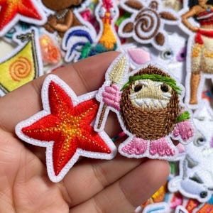 May include: Embroidered patches featuring characters and elements from the animated film "Moana." Includes a red starfish, a coconut character with a spear, and the film's title. Patches have white borders and are on a white background.