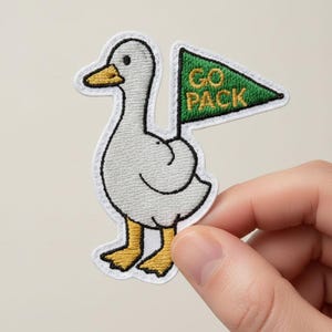 Op de afbeelding: Geborduurde patch met een witte eend met gele snavel en poten, die een groene vlag vasthoudt met de gouden tekst "GO PACK". De patch heeft een witte rand en wordt door een hand vastgehouden.