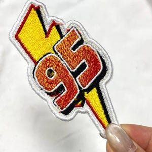 May include: Embroidered patch featuring the number "95" in orange and red, outlined in black, with a yellow lightning bolt. The patch has a white border and is likely for clothing or accessories.