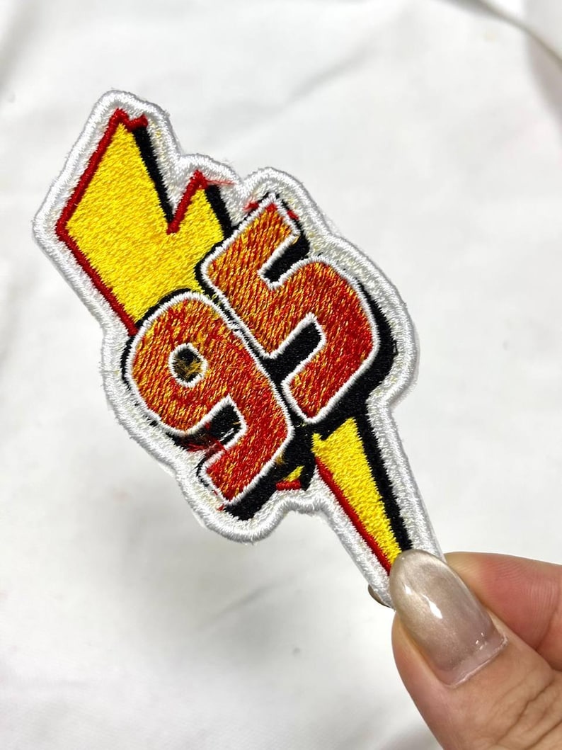May include: Embroidered patch featuring the number "95" in orange and red, outlined in black, with a yellow lightning bolt. The patch has a white border and is likely for clothing or accessories.