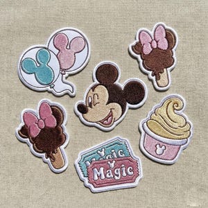 Puede incluir: Parches bordados con Mickey Mouse y diseños relacionados. Incluye la cara de Mickey, helados, una magdalena, globos y entradas con la palabra "Magic". Los parches tienen bordes blancos sobre un fondo beige.
