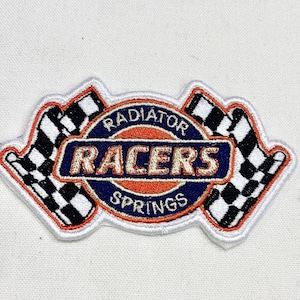 May include: Embroidered patch featuring the text "Radiator Racers Springs" in a circular design with a blue and orange color scheme. Two checkered flags flank the design, all outlined in white.