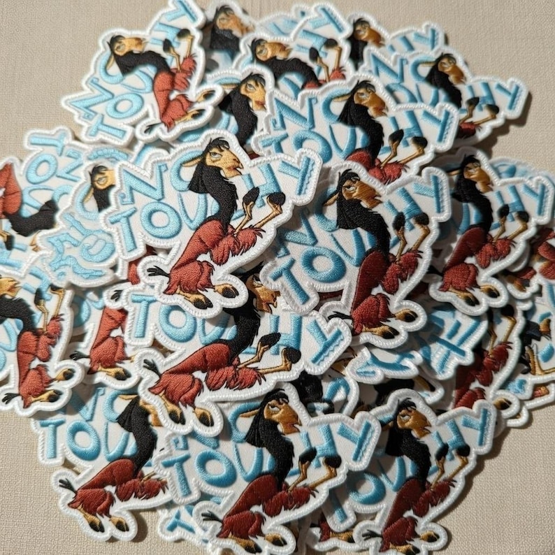 May include: A pile of embroidered patches featuring a cartoon llama character with a black mane and red fur. The patches have a white border and the words "NO TOUCHY" in blue letters.