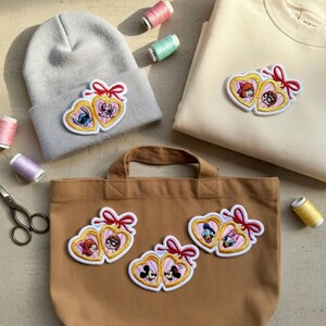 Op de afbeelding: Een lichtgrijze beanie, beige sweatshirt en bruine totebag zijn versierd met geborduurde hartvormige patches. Elke patch toont twee stripfiguren, een rode strik en een gouden rand. Naaibenodigdheden, waaronder garen en schaar, zijn verspreid.