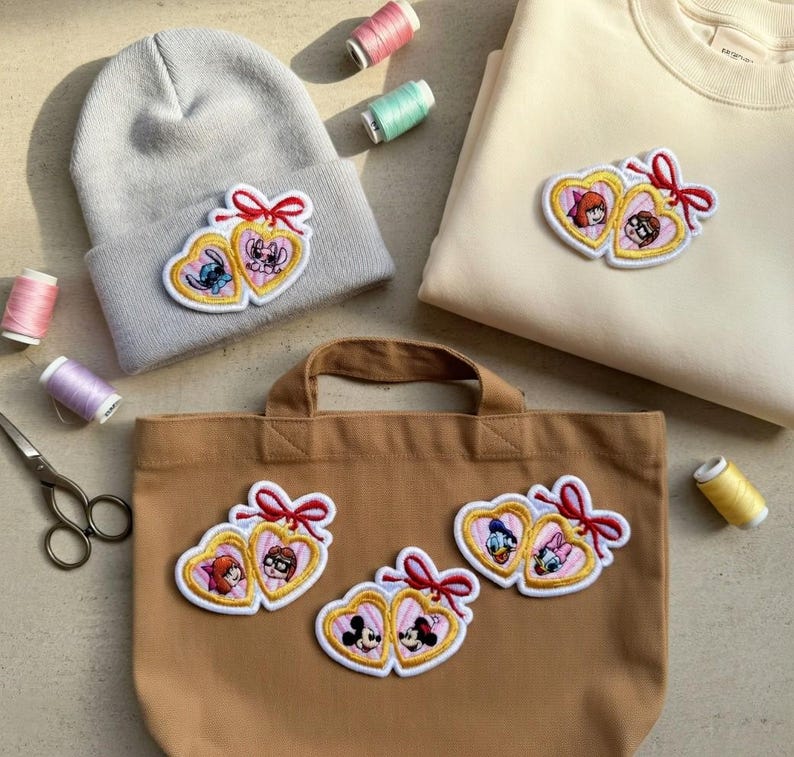 Op de afbeelding: Een lichtgrijze beanie, beige sweatshirt en bruine totebag zijn versierd met geborduurde hartvormige patches. Elke patch toont twee stripfiguren, een rode strik en een gouden rand. Naaibenodigdheden, waaronder garen en schaar, zijn verspreid.