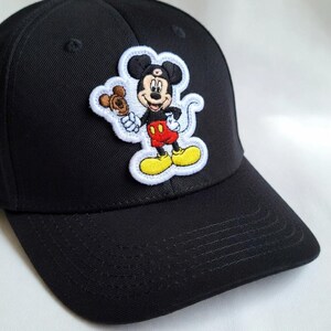 May include: Black baseball cap featuring an embroidered patch of Mickey Mouse holding a treat. The patch has a white border and showcases Mickey in his classic red shorts, yellow shoes, and black gloves. The cap is made of a durable fabric.