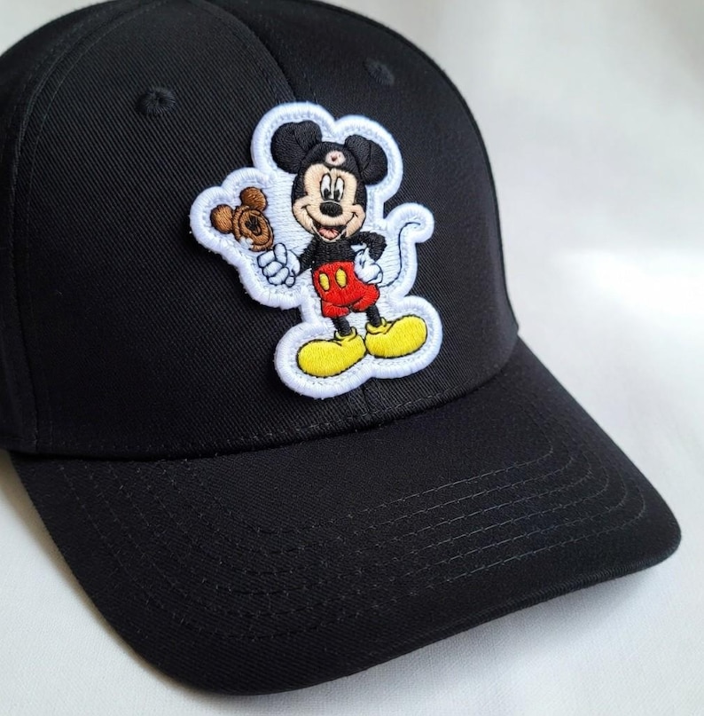 May include: Black baseball cap featuring an embroidered patch of Mickey Mouse holding a treat. The patch has a white border and showcases Mickey in his classic red shorts, yellow shoes, and black gloves. The cap is made of a durable fabric.