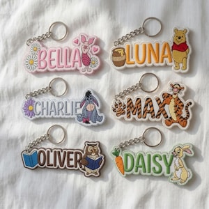 Könnte beinhalten: Sechs bestickte Schlüsselanhänger mit Zeichentrickfiguren und Namen. Die Anhänger zeigen die Namen Bella mit Ferkel, Luna mit Winnie Puuh, Charlie mit I-Aah, Max mit Tigger, Oliver mit Eule und Daisy mit Kaninchen, jeweils mit einem Metallring.