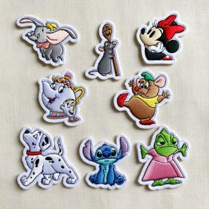 Op de afbeelding: Een verzameling geborduurde patches met verschillende stripfiguren. De patches tonen Dumbo, Minnie Mouse, een rat met een lepel, Chip, een Dalmatische hond, Stitch, een theepot en een groene kameleon in een roze jurk. Elke patch heeft een witte rand.