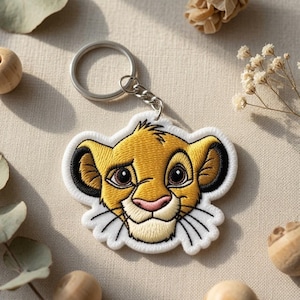 May include: A circular metal keyring attached to an embroidered patch featuring the face of a young lion cub. The lion has golden yellow fur, dark brown inner ears, large brown eyes, a pink nose, and white whiskers, all outlined in white thread.